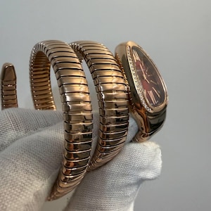 Puede incluir: Un reloj de oro rosa con una pulsera flexible en forma de serpiente enroscada. La esfera es ovalada con un fondo rojo oscuro, números romanos y un bisel engastado con pequeñas piedras claras. Los eslabones metálicos son segmentados y pulidos.