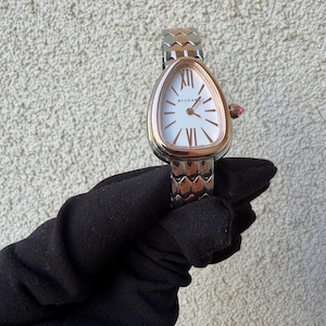 May include: A rose gold and silver wristwatch with a white face and Roman numeral hour markers. The watch has a unique teardrop-shaped face and a textured band. The watch is held by a gloved hand.