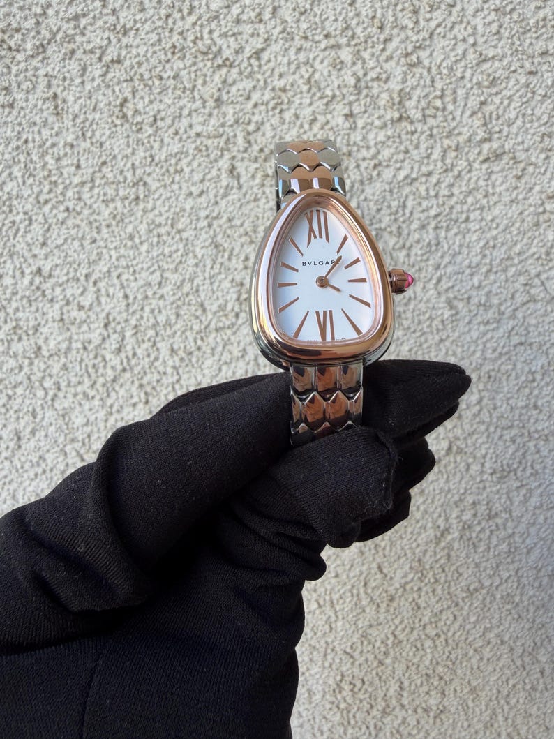 May include: A rose gold and silver wristwatch with a white face and Roman numeral hour markers. The watch has a unique teardrop-shaped face and a textured band. The watch is held by a gloved hand.