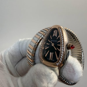 May include: A Bvlgari Serpenti watch featuring a teardrop-shaped black dial with rose gold hands and hour markers. The bezel is set with diamonds, and the watch includes a flexible two-tone rose gold and silver-coloured metal bracelet with a red cabochon crown.