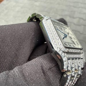 Könnte beinhalten: Eine silberfarbene Armbanduhr mit quadratischem Zifferblatt, römischen Ziffern und blauen Zeigern. Die Lünette und das integrierte Metallgliederarmband sind mit zahlreichen klaren Steinen besetzt. Auf dem Zifferblatt steht der Markenname Cartier.