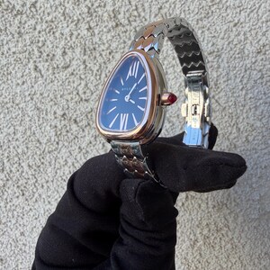 May include: A luxury wristwatch with a triangular face and a silver and rose gold band. The watch face is dark blue with white Roman numerals and hands. A small red gem accents the side of the watch. The watch is held against a textured gray background.