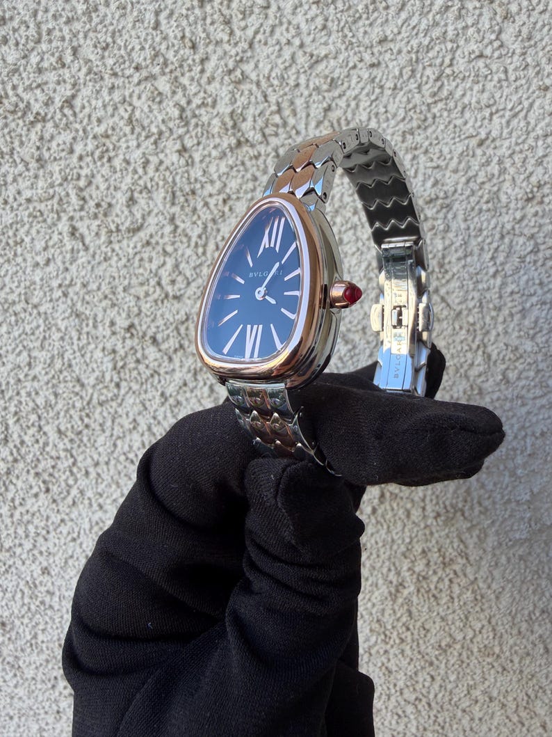 May include: A luxury wristwatch with a triangular face and a silver and rose gold band. The watch face is dark blue with white Roman numerals and hands. A small red gem accents the side of the watch. The watch is held against a textured gray background.