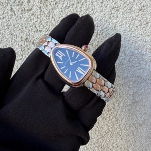May include: A luxury wristwatch with a blue face and gold-tone bezel. The watch has a snake-scale design band in silver and rose gold tones. The watch is held by a person wearing a black glove.