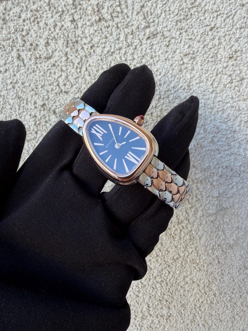 May include: A luxury wristwatch with a blue face and gold-tone bezel. The watch has a snake-scale design band in silver and rose gold tones. The watch is held by a person wearing a black glove.