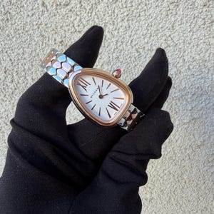 May include: A luxury wristwatch with a white face and gold-tone bezel. The watch features Roman numeral hour markers and a red gemstone on the crown. The watch band is silver and gold-tone with a geometric pattern.
