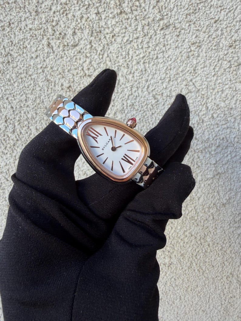 May include: A luxury wristwatch with a white face and gold-tone bezel. The watch features Roman numeral hour markers and a red gemstone on the crown. The watch band is silver and gold-tone with a geometric pattern.