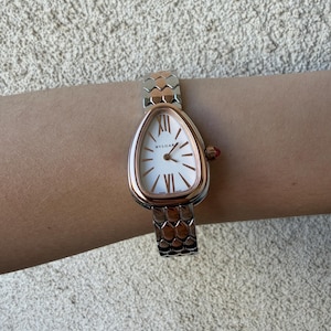 May include: A rose gold and silver wristwatch with a white oval face. The watch features Roman numerals and the brand name "BVLGARI". The watch band has a scale-like design. A small red jewel accents the side of the watch.