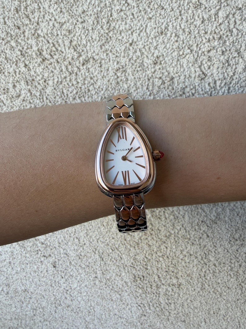 May include: A rose gold and silver wristwatch with a white oval face. The watch features Roman numerals and the brand name "BVLGARI". The watch band has a scale-like design. A small red jewel accents the side of the watch.