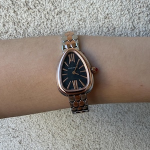 May include: A rose gold and silver wristwatch with a black face and Roman numeral hour markers. The watch has a snake-like design on the band and a red crown. The brand name "BVLGARI" is visible on the watch face.
