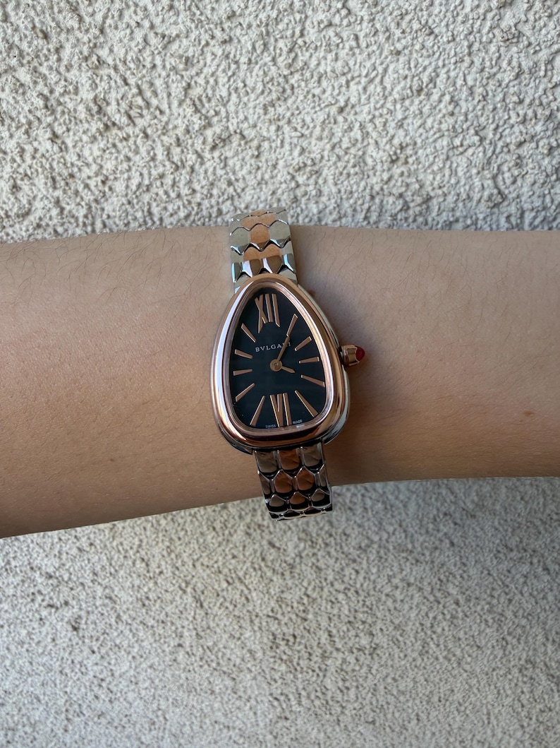 May include: A rose gold and silver wristwatch with a black face and Roman numeral hour markers. The watch has a snake-like design on the band and a red crown. The brand name "BVLGARI" is visible on the watch face.