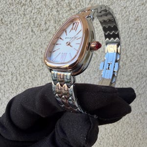 May include: A luxury wristwatch with a white face and rose gold bezel. The watch features Roman numeral hour markers and the brand name "BVLGARI". The silver and rose gold band is connected to the watch with a red gem. The watch is held by a black gloved hand.