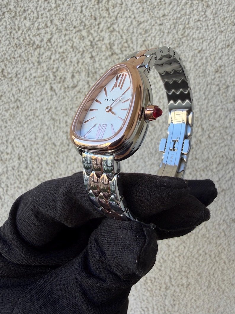May include: A luxury wristwatch with a white face and rose gold bezel. The watch features Roman numeral hour markers and the brand name "BVLGARI". The silver and rose gold band is connected to the watch with a red gem. The watch is held by a black gloved hand.