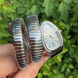 Puede incluir: Un reloj Bulgari Serpenti de tono plateado con una distintiva pulsera tubogas envolvente de diseño segmentado. El reloj tiene una caja en forma de gota con esfera plateada, marcadores de números romanos y un bisel decorado con piedras claras.