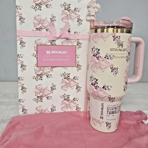Könnte beinhalten: Ein cremefarbener Stanley Quencher-Becher mit Blumen- und Schleifenmuster, einem rosa Griff und einer passenden Geschenkbox. Auf der Box und dem Becher steht der Text "LoveShackFancy". Der Becher hat ein Fassungsvermögen von 1,18 Litern.