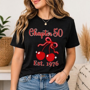 Puede incluir: Camiseta negra con el texto "Chapter 50" en escritura roja sobre un par de cerezas rojas atadas con un lazo rosa. Debajo de las cerezas está "Est. 1976" en rojo. La camiseta está siendo usada.