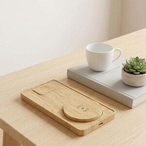 Pode incluir: Uma estação de carregamento sem fio de bambu com uma almofada de carregamento circular e uma base retangular. Uma caneca de cerâmica branca, uma planta suculenta em um vaso pequeno e um livro estão em uma mesa de madeira clara.