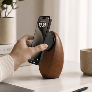 May include: A dark wood wireless charging stand with a smooth, rounded, pebble-like shape. A black smartphone is magnetically attached to the circular charging pad on the front of the wooden base, displaying the time 10:30 on its screen.