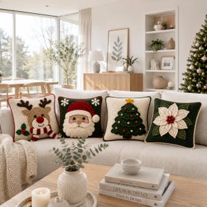 May include: Four square crochet throw pillows arranged on a sofa. The first features a reindeer with a red nose, the second a Santa Claus face, the third a decorated Christmas tree, and the fourth a white poinsettia flower on a dark green background.