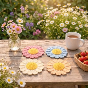 Może przedstawiać: Cztery ręcznie robione podkładki w kształcie stokrotek w kolorze różowym, niebieskim, kremowym i brzoskwiniowym z żółtymi środkami leżą na drewnianym stole. Na stole stoi również biały kubek z napojem, mały wazon z kwiatami i kosz truskawek.