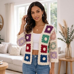 Puede incluir: Un cárdigan de crochet abierto y colorido. La chaqueta presenta paneles cuadrados en crema, morado, verde, rojo y azul, con un borde crema festoneado. Las mangas son de tres cuartos. La mujer lleva un top blanco y vaqueros azules.