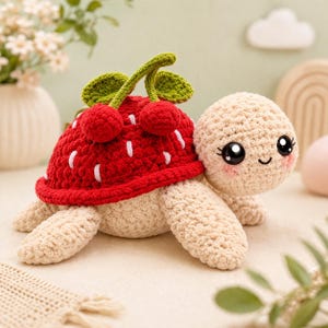 May include: A crocheted turtle plushie with a red strawberry shell and green leaves. The turtle has a beige body, legs, and head, with large black eyes and rosy cheeks. The strawberry shell has white seed details.