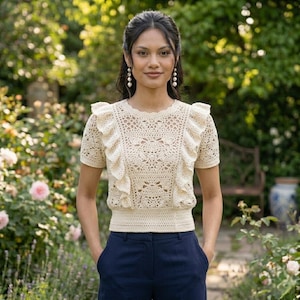 May include: Cream-colored crochet top with short sleeves and ruffled accents. The top features an intricate openwork design and is paired with navy blue pants. The image is set outdoors with a garden background.