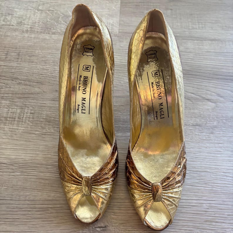 May include: A pair of gold-colored high heels with a textured, snakeskin-like pattern. The shoes have a peep-toe design and a bow detail on the front. The brand name "Bruno Magli" is visible inside the shoe.