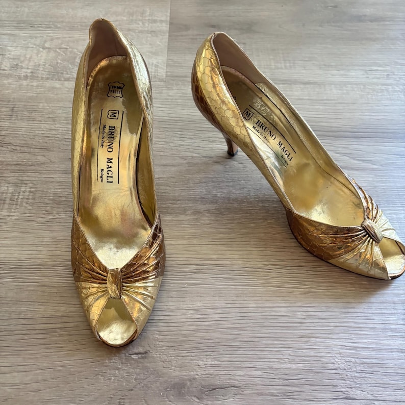 May include: Pair of gold-colored peep-toe high heels. The shoes have a textured, snakeskin-like pattern and a bow detail on the toe. The brand name "BRUNO MAGLI" is visible inside the shoe.