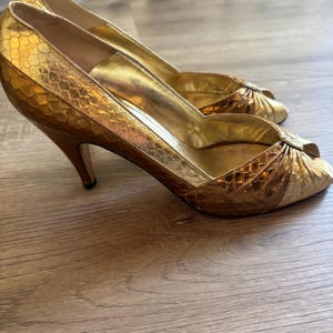 May include: A pair of gold high-heeled shoes with a snakeskin pattern. The open-toe heels have a stiletto heel and a bow detail on the front. The shoes are made of a shiny, metallic material.