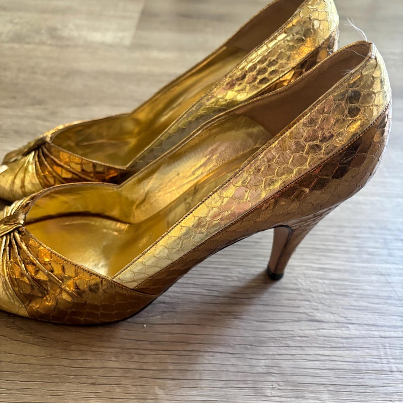 May include: A pair of gold high heels with a textured, snakeskin-like pattern. The shoes have a pointed toe and a stiletto heel. The interior of the shoe is a solid gold color.