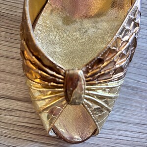 May include: Close-up of a gold-colored shoe with a bow detail. The shoe has a textured, snakeskin-like pattern and a metallic sheen. The interior is a lighter gold color.