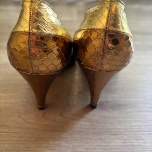 May include: A pair of gold-colored high heels with a textured, scale-like pattern. The heels have a pointed toe and a slender, curved heel. The shoes are made of a shiny material and are set against a wooden surface.
