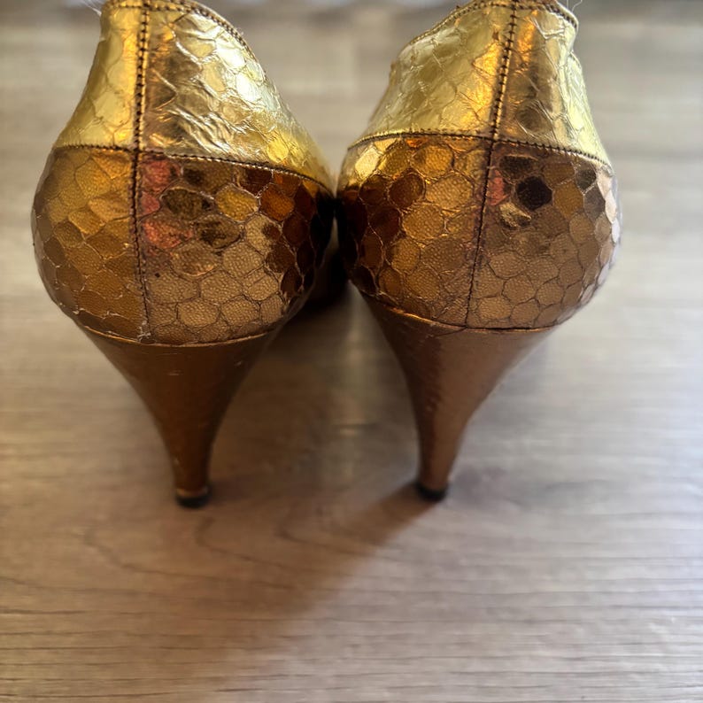 May include: A pair of gold-colored high heels with a textured, scale-like pattern. The heels have a pointed toe and a slender, curved heel. The shoes are made of a shiny material and are set against a wooden surface.