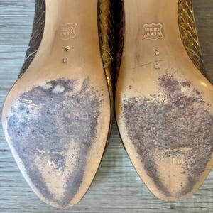 May include: Pair of used, gold-toned high heels with a snakeskin pattern. The soles show wear and tear, with visible scuff marks and discoloration. The heels are size 9 B, and the soles are marked "VERO CUOIO".