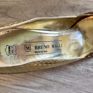 May include: Close-up of the interior of a gold-colored shoe. The shoe's insole features the brand name "BRUNO MAGLI" and "Made in Italy". The shoe also has a leather symbol and the word "TOMMA PELLE".