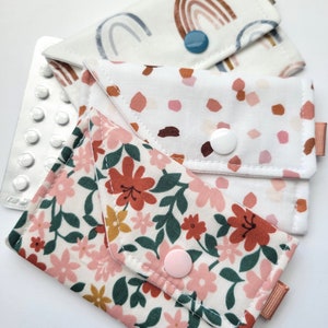 May include: Three fabric pouches with different patterns. The first pouch has a white background with a rainbow pattern in blue and brown. The second pouch has a white background with brown and pink polka dots. The third pouch has a white background with a floral pattern in pink, red, green, and yellow.