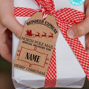 Puede incluir: Una caja de regalo blanca con una cinta a cuadros rojos y blancos. Una etiqueta de regalo marrón con el texto "Reindeer Express North Pole Mail Special Delivery For NAME Merry Christmas" está unida a la cinta.
