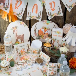 May include: A woodland themed baby shower party set with a fawn and fox design. The set includes plates, cups, napkins, favors, and decorations. The colors are orange, brown, and green.