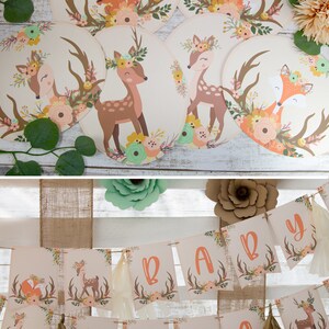 May include: A woodland themed baby shower banner with the word "BABY" in large letters. The banner is decorated with floral patterns, antlers, and deer silhouettes. The banner is strung on a rustic twine.