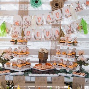 May include: A woodland themed baby shower dessert table with cupcakes decorated with deer and fox designs. The table is decorated with a banner that says "Baby Nora" and has paper flowers in shades of green, orange, and brown.