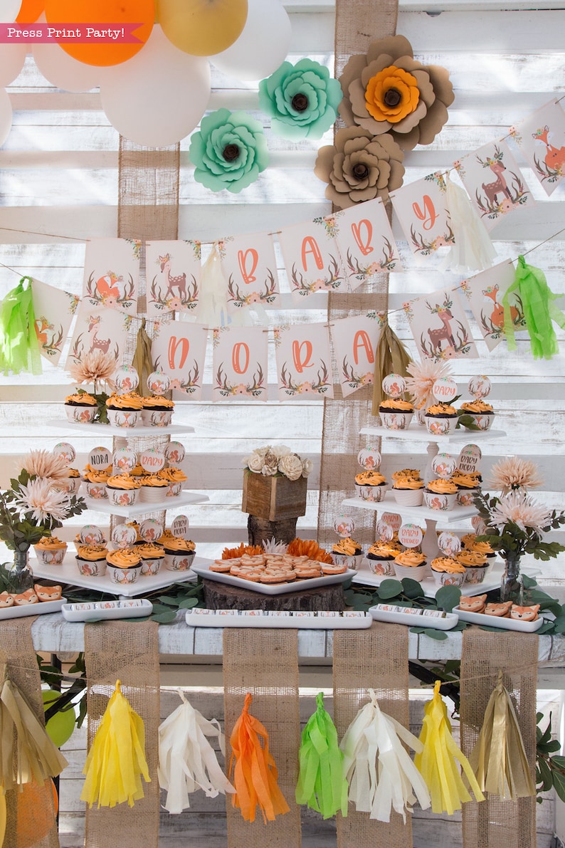 May include: A woodland themed baby shower dessert table with cupcakes decorated with deer and fox designs. The table is decorated with a banner that says "Baby Nora" and has paper flowers in shades of green, orange, and brown.