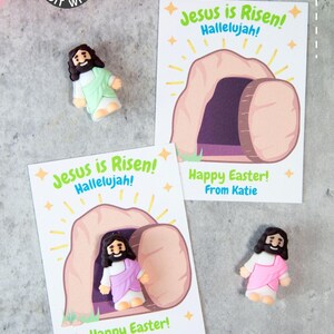 Pode incluir: Cart&otilde;es de felicita&ccedil;&otilde;es de P&aacute;scoa com o texto "Jesus ressuscitou! Aleluia!" e "Feliz P&aacute;scoa!" juntamente com pequenas figuras de Jesus. Os cart&otilde;es apresentam uma ilustra&ccedil;&atilde;o do t&uacute;mulo vazio. O fundo inclui papel rosa.