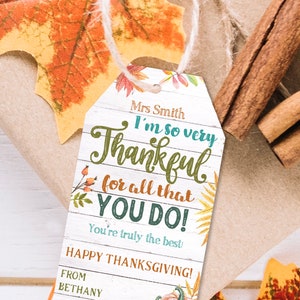 May include: A printable Thanksgiving gift tag with a white woodgrain background. The tag features a handwritten message that says "I'm so very Thankful for all that YOU DO! You're truly the best! Happy Thanksgiving!" with fall-themed illustrations. The text is editable except for the word "Thankful".