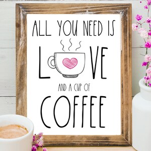 K&ouml;nnte beinhalten: Ein gerahmter Druck mit dem Text "Alles was du brauchst ist Liebe und eine Tasse Kaffee" in schwarzer Schrift. Eine stilisierte Zeichnung einer Kaffeetasse mit einem rosa Herzen darin befindet sich zwischen den W&ouml;rtern "Liebe" und "und".
