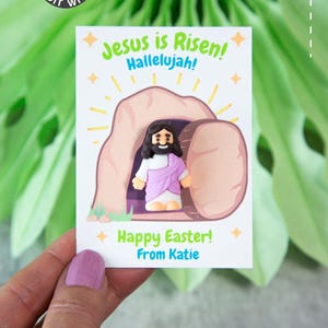 May include: A white card with the text "Jesus is Risen! Hallelujah!" and "Happy Easter! From Katie." An illustration of Jesus emerging from a tomb is featured. The card is held against a green background.