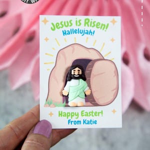 Pode incluir: Um cart&atilde;o branco com uma ilustra&ccedil;&atilde;o de desenho animado de Jesus saindo de um t&uacute;mulo, com o texto "Jesus ressuscitou! Aleluia!" e "Feliz P&aacute;scoa!" em verde e azul. O cart&atilde;o &eacute; segurado em uma m&atilde;o.