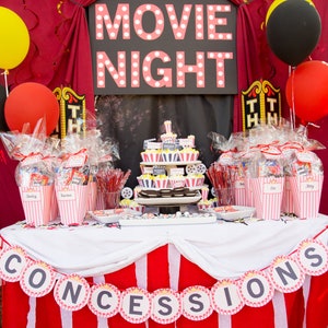 Op de afbeelding: Een feesttafel met een filmnacht thema met een rood-wit gestreept tafelkleed. De tafel is versierd met een banner met de tekst "CONCESSIONS" en een bord met de tekst "FILMAVOND". Er zijn cupcakes met popcorn en filmroldecoraties en feestcadeaus in gestreepte zakken.