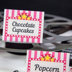 May include: Two pink and white striped signs with a marquee design and gold stars. The top sign says "Chocolate Cupcakes" and the bottom sign says "Popcorn".