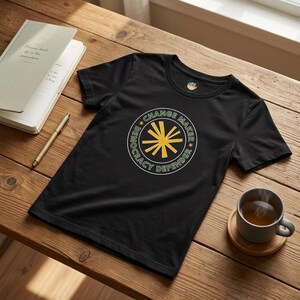 May include: Black t-shirt with a circular graphic that reads "Democracy Defender Change Maker" around a yellow starburst design. The shirt is displayed on a wooden surface with a notebook, pen, and a cup of tea.